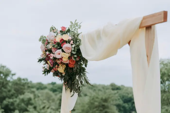 A wooden ceremony arch draped with fabric and flowers, facing open countryside.