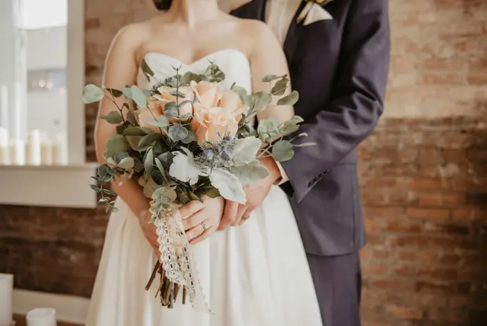 A bride holding a bouquet in front of a brick wall, next to the groom.