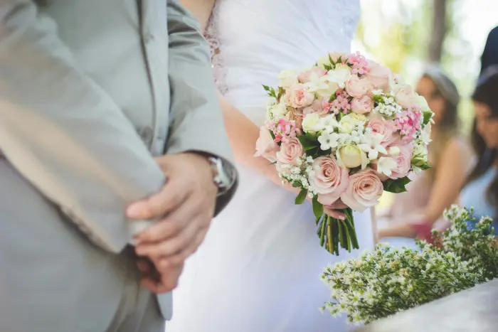 A groom's hand holding the bride's hand and her bouquet of pink roses.
