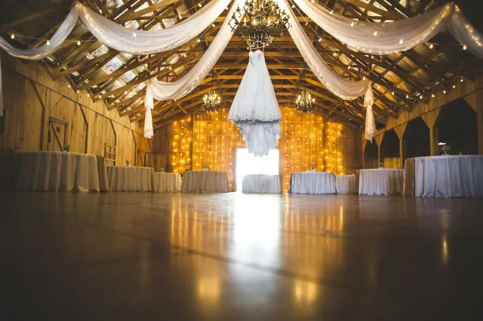 The inside of a barn lit warmly, with a chandelier hanging from the wooden beams and fabric draped across the ceiling.