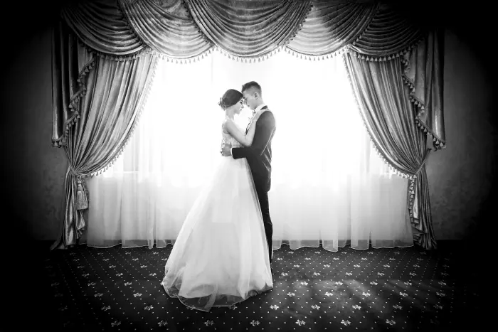 A black and white photo of a couple embracing in front of tall curtains.