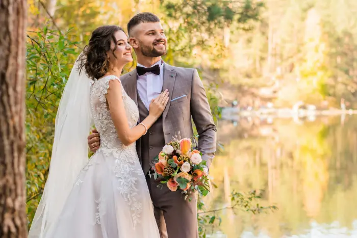 A couple smiling by a lake, the groom holding the bouquet.
