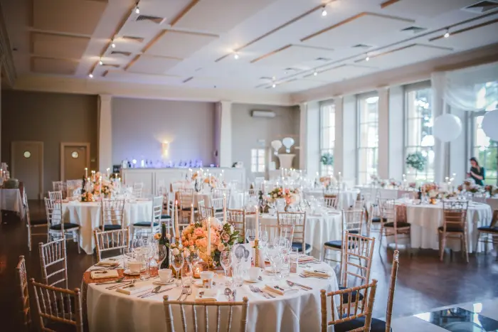 An elegant reception room with round tables, white linens, chandeliers and tall windows.